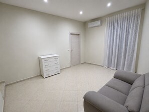 A room with a grey sofa, a white dresser, a closed door, and a window with white curtains.