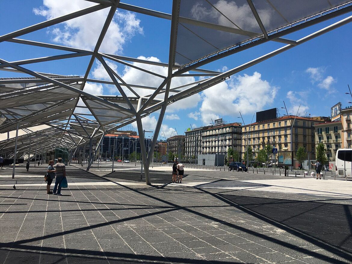 A covered pedestrian area with a geometric metal framework, a paved walkway, and buildings in the background.