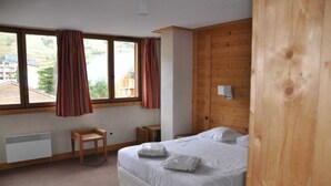 Une chambre avec un grand lit, une tête de lit en bois, une petite table de chevet en bois, une fenêtre avec des rideaux et une vue sur les bâtiments à l’extérieur.