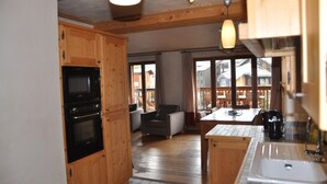 Une cuisine avec des meubles en bois, un four noir et un évier blanc. On y trouve un coin repas avec une table et des chaises, ainsi qu’une fenêtre avec des rideaux.
