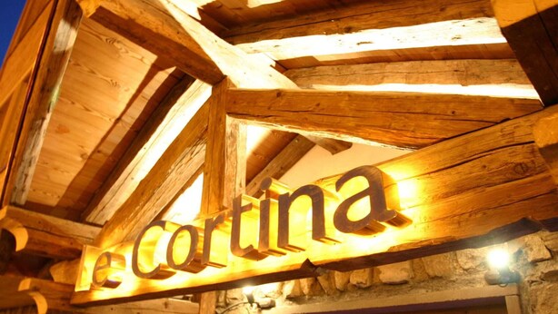 Une enseigne en bois portant le nom « Le Cortina » est illuminée sur un mur de pierre.