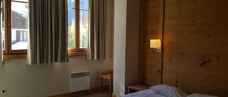A bedroom with a wooden headboard, a bed with white linens, a window with curtains, a chair, and a wall-mounted lamp.