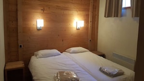 Ett rum med träpanel på väggarna, med två sängar, båda med vita sängkläder och kuddar. Det finns två väggmonterade lampor och ett fönster med gardiner.