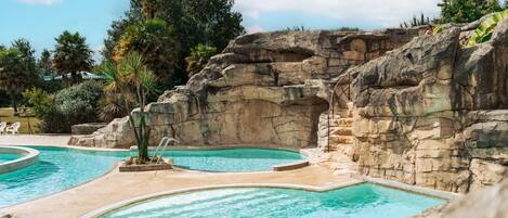 Un espace piscine comprenant plusieurs piscines, un décor rocheux et une végétation luxuriante tout autour.