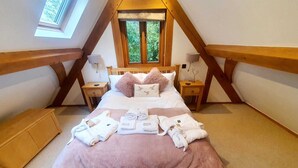 Une chambre avec un plafond en pente, un lit recouvert d’une couverture rose, deux tables de chevet avec des lampes, une fenêtre donnant sur la verdure et un petit banc en bois.