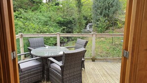 Un balcon extérieur aménagé avec des meubles en osier et une table en verre, offrant une vue sur un jardin luxuriant et une petite cascade.