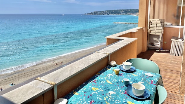 Un balcón con una mesa para dos, con vista a una playa de aguas azules cristalinas y una costa distante.