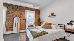 Ein modernes Schlafzimmer mit Backsteinwand, einem großen Bett mit weißer Bettwäsche, einem kleinen Nachttisch mit Lampe und einem Fenster mit Vorhängen.