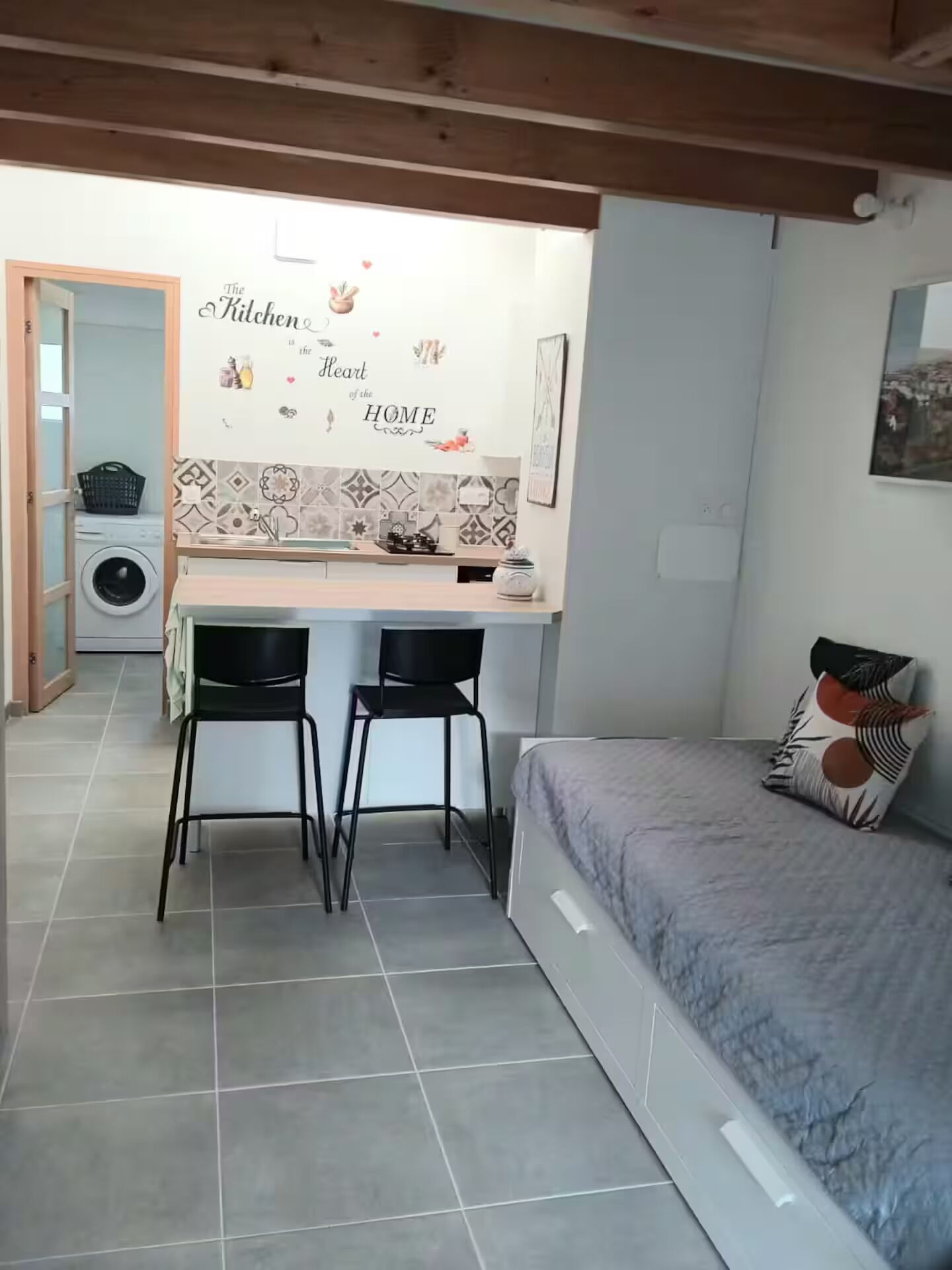 Une cuisine compacte avec un crédence carrelée, un coin repas avec deux chaises et un lit agrémenté d’oreillers à motifs.