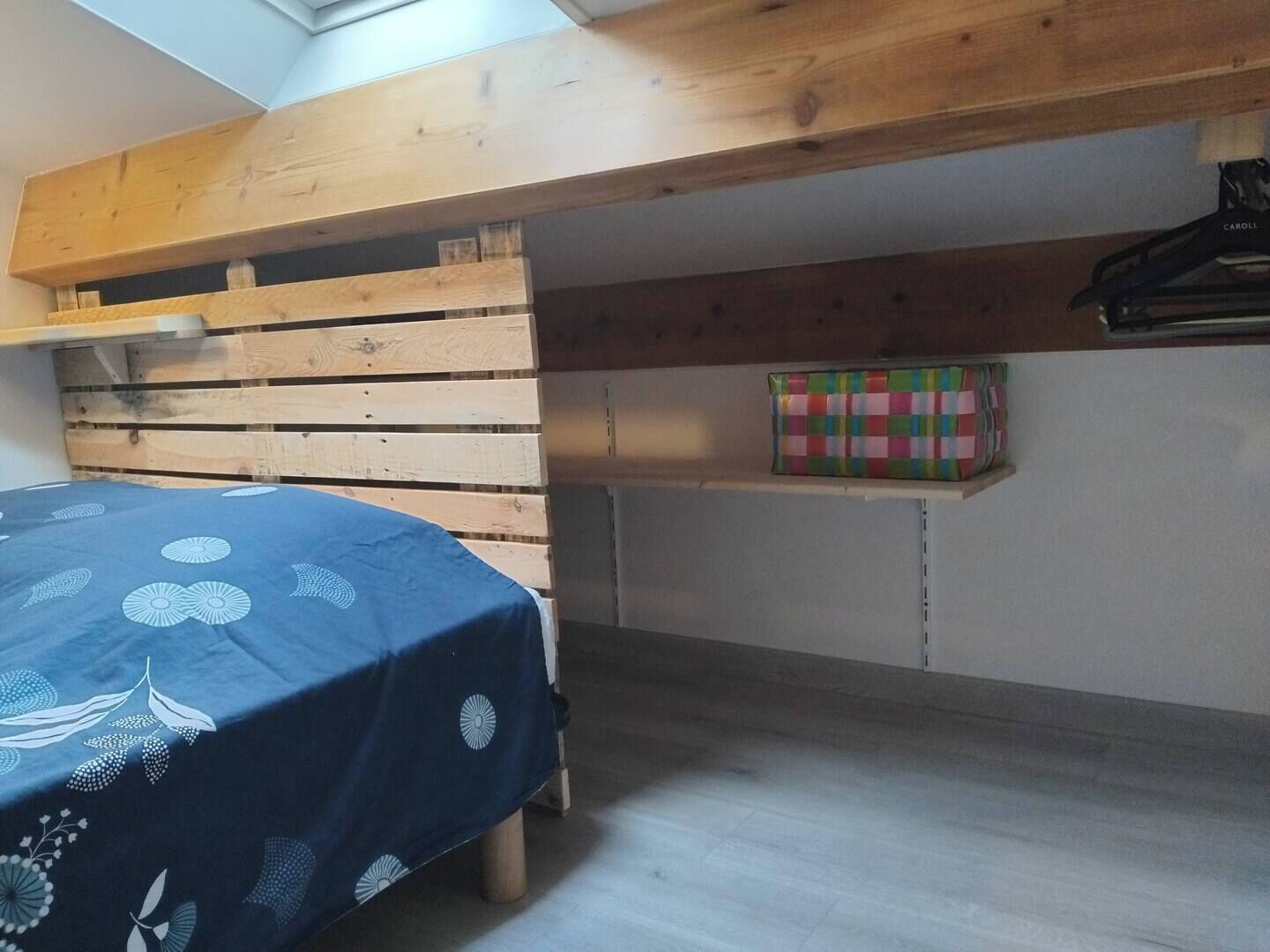 Un dormitorio con cama, cabecera de madera, repisa con una caja colorida y una prenda colgando.