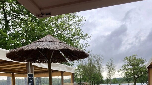 An outdoor seating area with wooden tables and benches, a thatched umbrella, and a view of a lake.