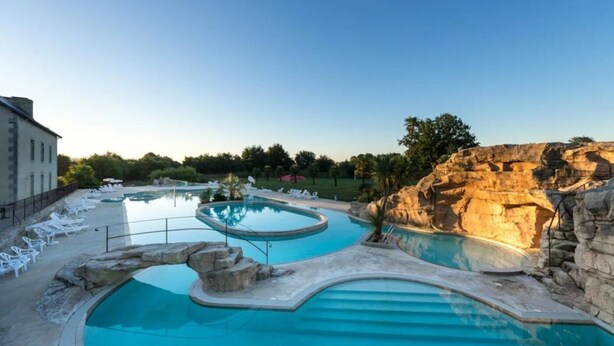 A swimming pool with lounge chairs, a rock formation, and a building in the background. - ZEcamping 5* parc aquatique club enfant fitness locatif BBQ 2 pers (Quimper)