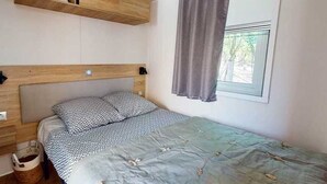 A bedroom with a wooden headboard, a bed with patterned bedding, a window with curtains, and a small shelf above the bed.