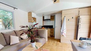 A modern living room with a sofa, a coffee table, and a potted plant. There is a kitchen area with cabinets and a refrigerator.