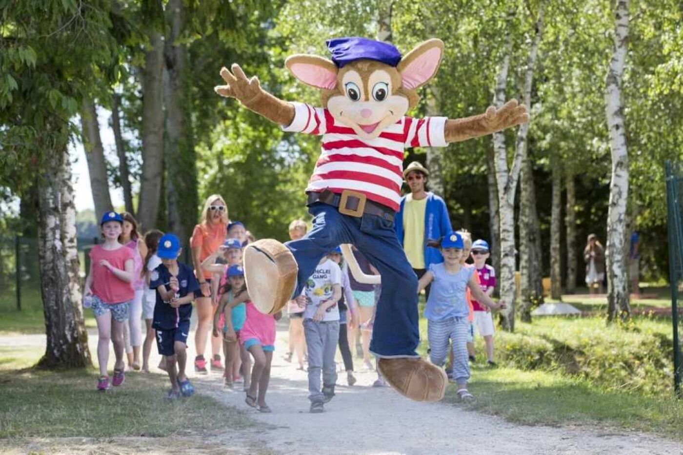 Un personnage mascotte, vêtu d’un maillot rayé et d’une casquette bleue, saute dans un parc où l’on aperçoit des arbres et des personnes au loin.