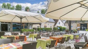 Ein Außenbereich mit Terrasse, Tischen und Stühlen, beschattet durch große Sonnenschirme, mit Steinplattenboden.