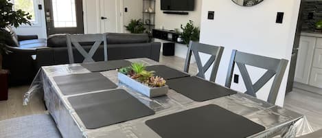 Ein modernes Wohnzimmer mit einer Couch, einem Esstisch mit schwarzen Tischsets, einem an der Wand montierten Fernseher und einem großen Spiegel.
