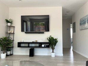 A modern living room with a flat-screen TV mounted on the wall, a black entertainment unit below, and two potted plants. - Oasis 2BR Culver City: 6 Miles to SoFi Stadium (Culver City)