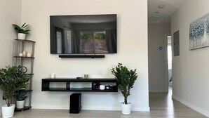 A modern living room with a large flat-screen TV mounted on the wall, a black entertainment unit below, and two potted plants on either side. - Cheerful house with EV, Parking & backyard (Culver City)