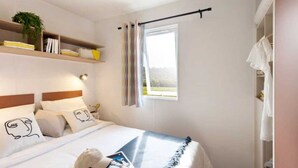 Une chambre à coucher comprenant un lit, des oreillers, une table de chevet, une fenêtre avec des rideaux, une étagère avec des livres et une plante, ainsi qu’une armoire.