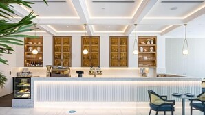 Un moderno vestíbulo de hotel con un mostrador de recepción central, vitrinas de vidrio y un área de descanso con sillas y una mesa.