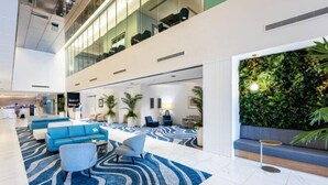 Un moderno vestíbulo de hotel con techo alto, alfombra estampada en azul y blanco, zonas de estar con sofás azules y una pared verde con plantas.