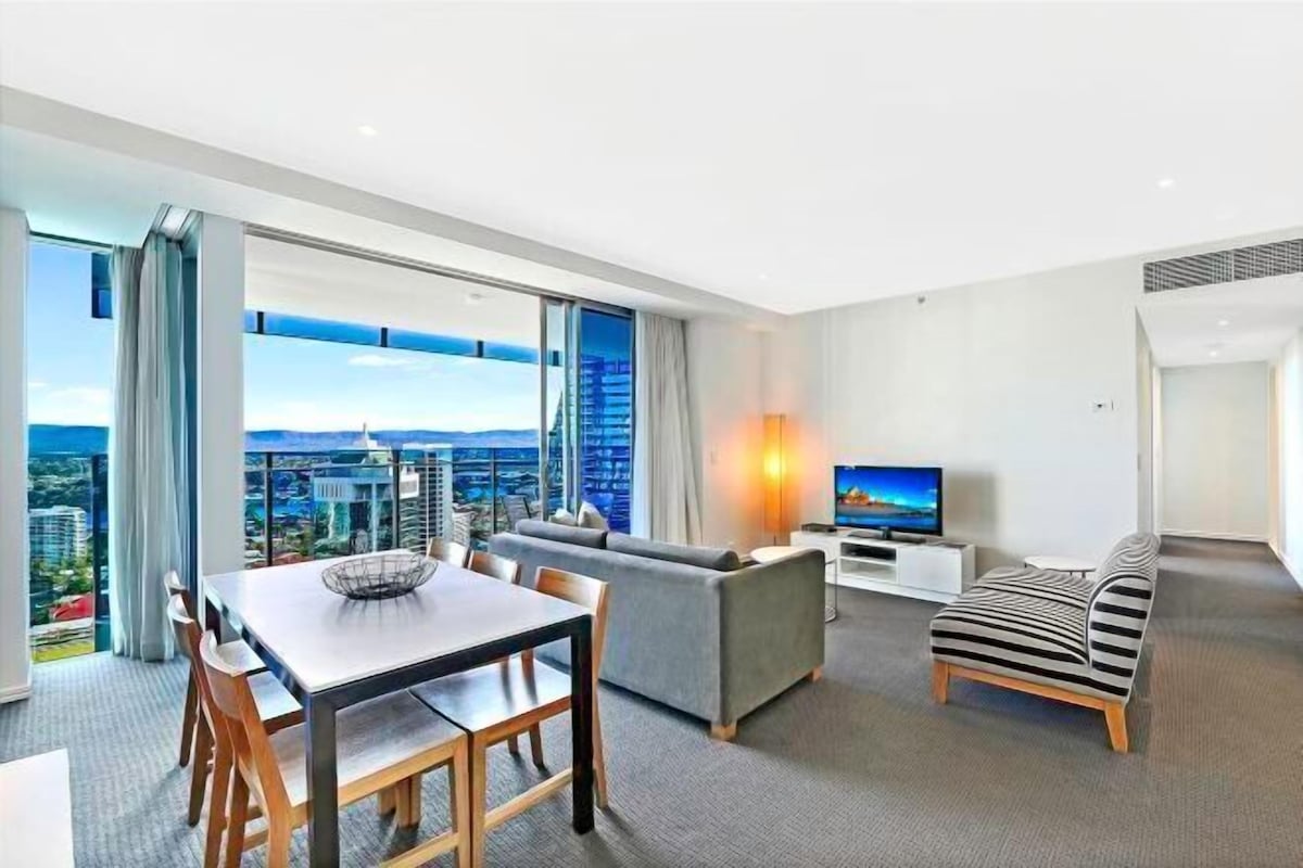 A modern hotel room with a dining area, sofa, TV, and a city view.