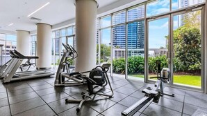 Un gimnasio con equipamiento de ejercicio, ventanas amplias y vistas a edificios y áreas verdes.