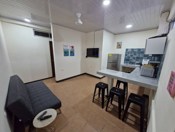 Una cocina compacta con refrigerador, microondas y una pequeña barra. Hay un banco con un control remoto, y un televisor montado en la pared.