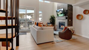 A spacious living room with a white sofa, a brown armchair, and a fireplace. A TV is mounted on the wall above the fireplace.