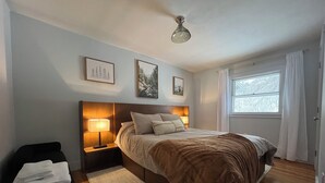 A bedroom with a bed, bedside lamps, a nightstand, a window with curtains, and artwork on the wall. - Enjoyable a cabin on the Black river with WiFi in Ludlow for a cozy stay (Ludlow)