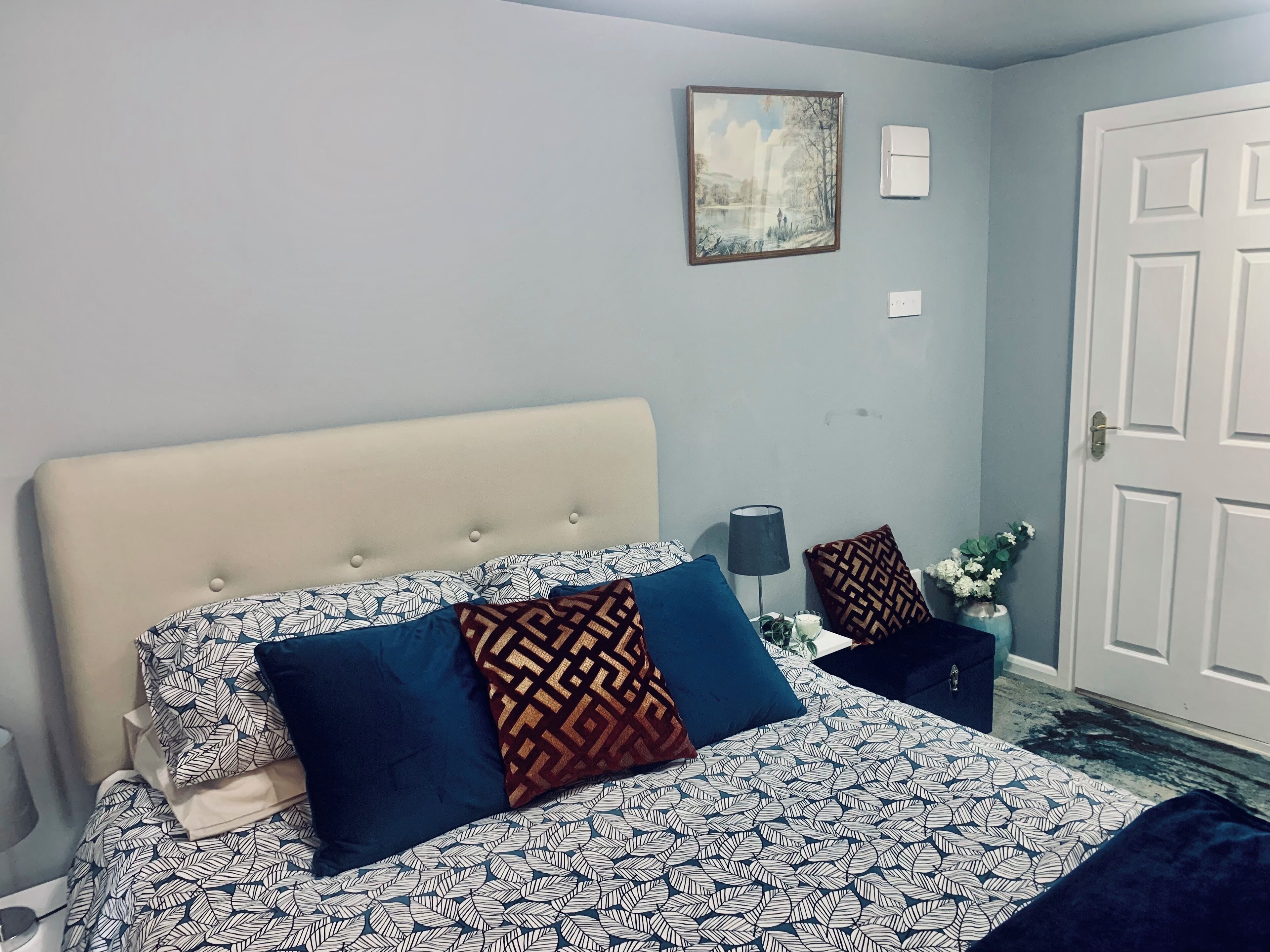 A neatly made bed with patterned bedding and decorative pillows, a bedside table with a lamp, a wall-mounted air conditioner, and a door with a simple design.