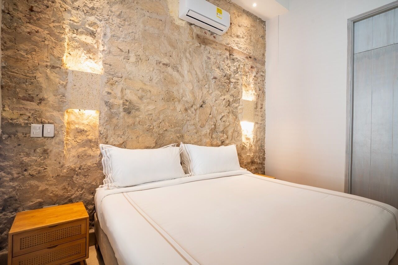 A bedroom with a stone wall, a bed with white linens, a wooden nightstand, and an air conditioning unit.