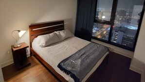 Un dormitori amb un llit de fusta, una tauleta de nit amb una làmpada, un finestral gran amb vista a la ciutat i una manta amb dibuixos.