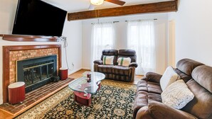 Una sala de estar con chimenea, televisor, ventilador de techo y un ventanal con cortinas.