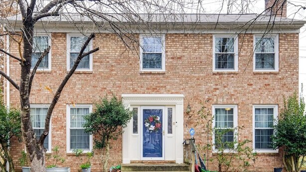 Una casa de ladrillo de dos pisos con puerta principal, varias ventanas y un pequeño jardín con plantas y un árbol.