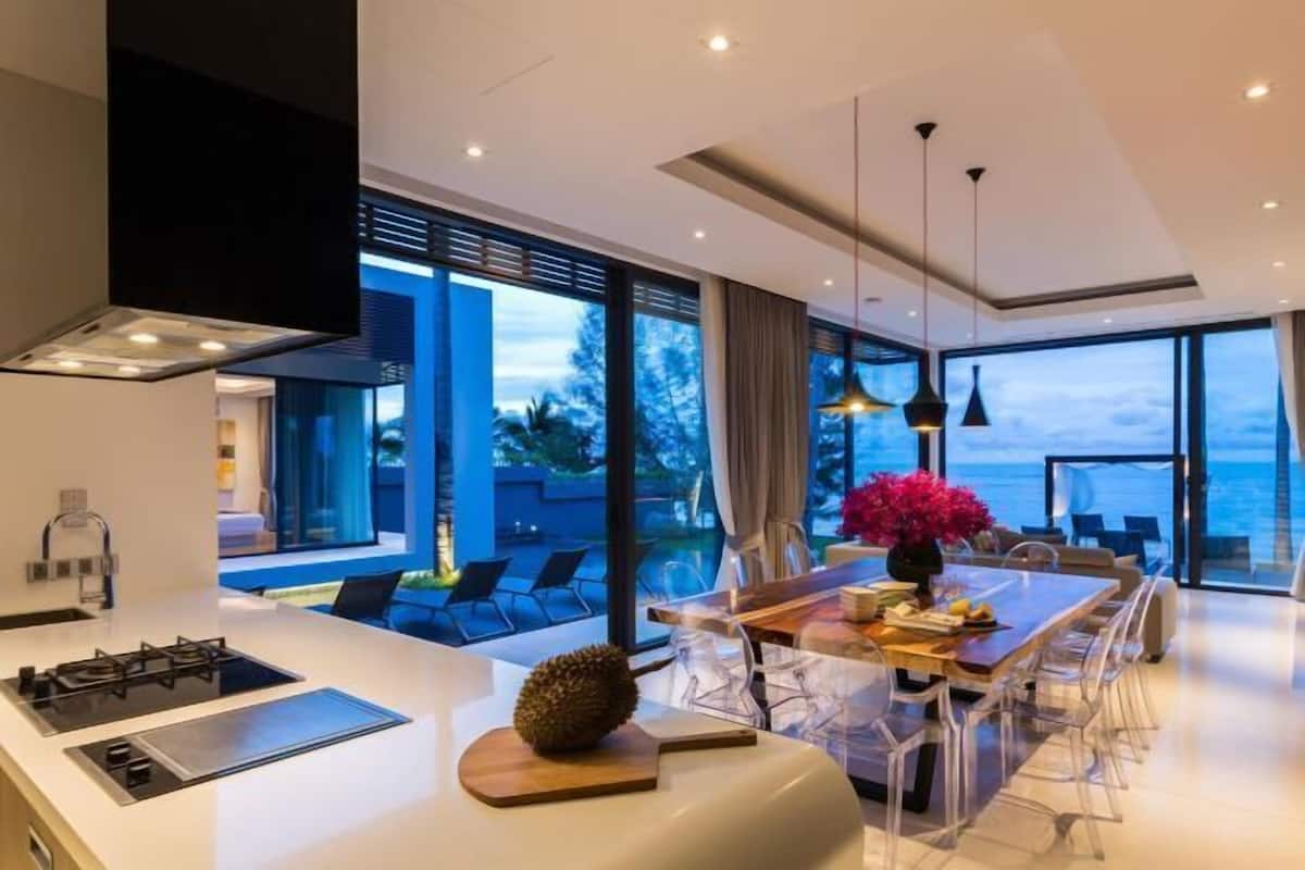A modern kitchen with a gas stove, a dining area with a wooden table and chairs, and large windows offering a view of the ocean.