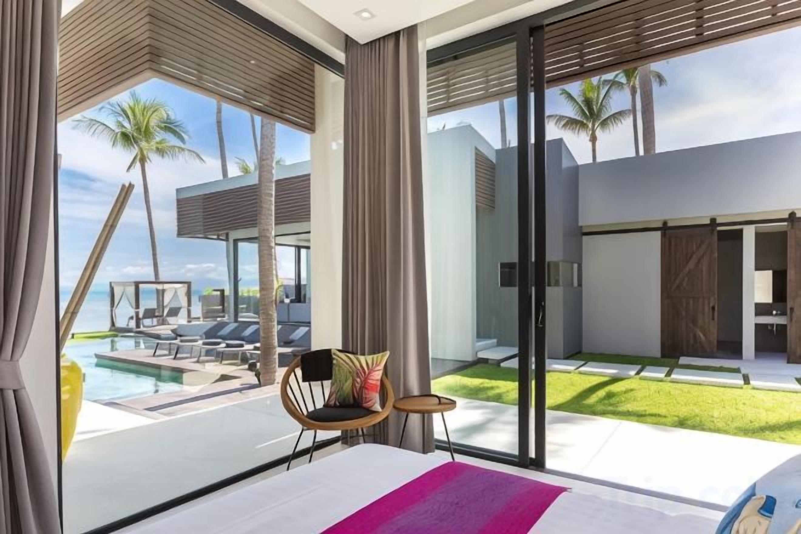 A modern hotel room with a large sliding glass door leading to a pool area, a bed with a colorful throw, and a chair with a cushion.