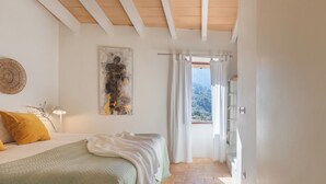 A bedroom with a bed, a painting, a lamp, and a view of a mountainous landscape through a window. - Can Palou Fornalutx (Fornalutx)
