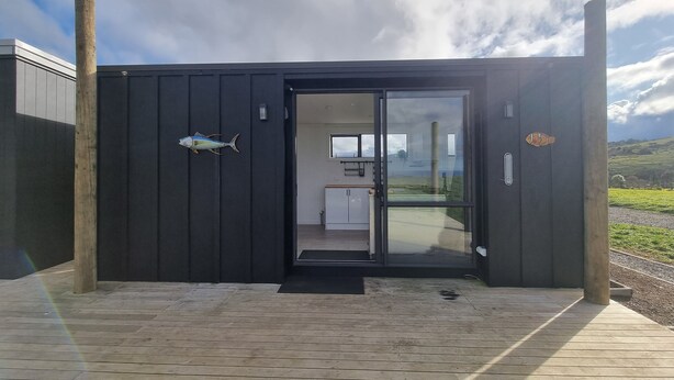 A modern building with a wooden deck entrance, a fish decoration, and a view of a kitchen and landscape through a glass door. - Marvelous cabin with AC in Helena Bay (Helena Bay)
