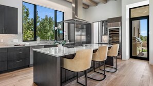 Una cocina moderna con una isla grande, armarios oscuros, electrodomésticos de acero inoxidable y vistas al exterior.
