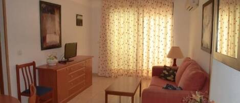 Una sala de estar con sofá, mesa de comedor, televisor en un mueble y ventana con cortinas.