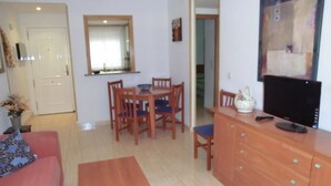 Ein Wohnzimmer mit einer Couch, einem Holztisch, einem Essbereich mit Tisch und Stühlen, einem Fernseher auf einer Holzkommode und einer Tür zu einem anderen Raum.