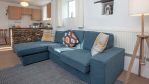 A modern living room with a blue sectional sofa, a patterned rug, a stone accent wall, and a kitchen in the background. - 2 bedroom accommodation in Looe (Looe)