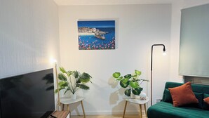 Un salon moderne doté d’un téléviseur à écran plat, de deux plantes en pot, d’un canapé vert agrémenté de coussins orange et d’un tableau représentant un paysage côtier.