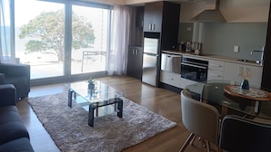 A modern kitchen with a large window, a dining area with a glass table, and a sofa. - Paihia Beach Front 1 bedroom Apartment (Paihia)
