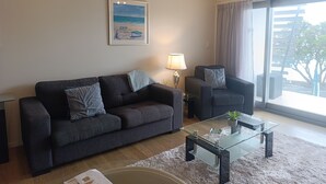 A living room with a dark grey sofa, a glass coffee table, and a painting of a beach scene. - Paihia Beach Front 1 bedroom Apartment (Paihia)