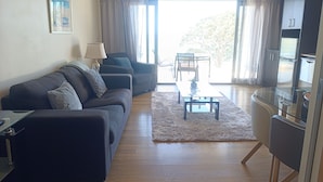 A modern living room with a sofa, a coffee table, and a dining area with chairs. There is a kitchen area with cabinets and a refrigerator. - Paihia Beach Front 1 bedroom Apartment (Paihia)