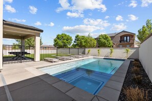 A rectangular swimming pool with a clear blue water surface, surrounded by a concrete deck and a metal fence. There are lounge chairs and a covered seating area with a table. - Brand New Build! 50+ Beds (Hurricane)