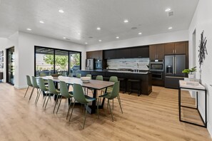 A modern kitchen with dark cabinets, a central island, and a dining area with green chairs. - Brand New Build! 50+ Beds (Hurricane)